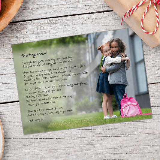 A printed personalized starting school landscape ard lies on a weathered wood surface, styled with a wrapped gift box, macarons, and a plate in the corners. The card features a photo of two girls hugging and a pink backpack, with a poem printed on the left side.