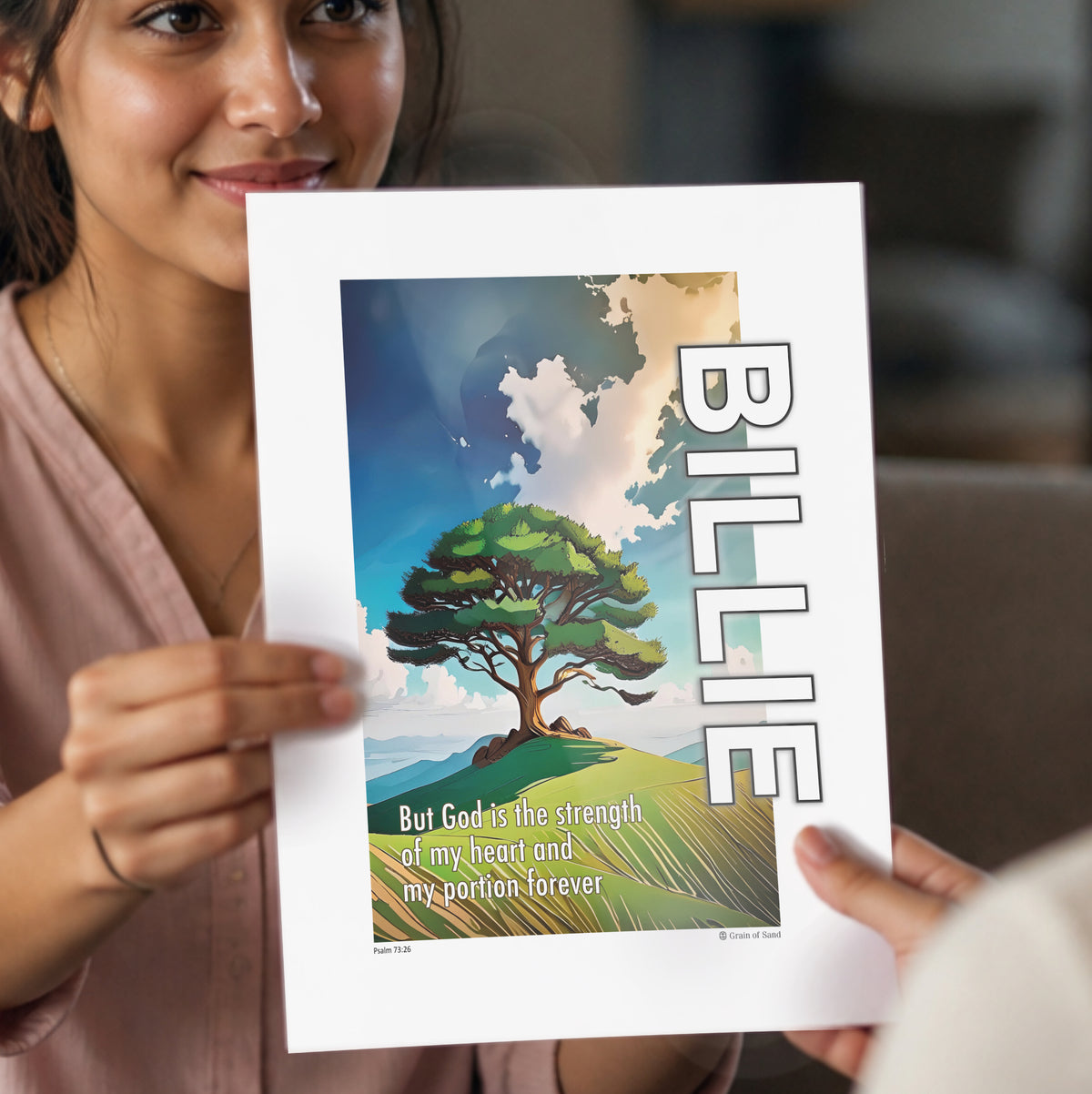 Woman holding a personalised A4 poster featuring a stylised tree on a hill with blue sky and clouds with the name Billie vertically up the side of the poster image and a biblical scripture Psalm 73 verse 26.