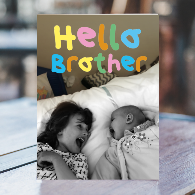 A custom photo print card featuring the text 'Hello Brother' in colorful bubble letters above a black-and-white photo of a baby and an older sister, displayed on a wooden surface with a blurred kitchen background.