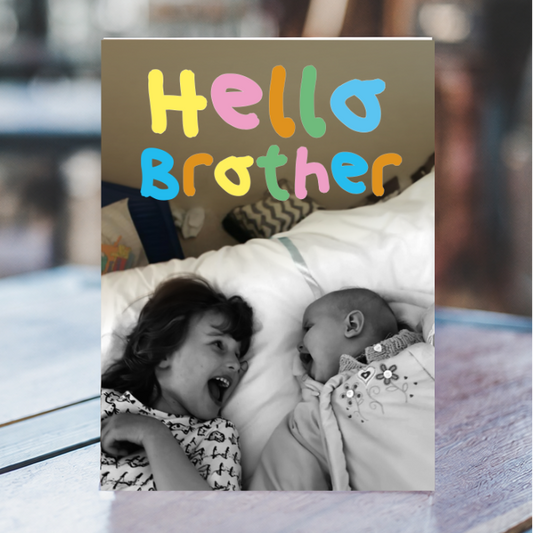 A custom photo print card featuring the text 'Hello Brother' in colorful bubble letters above a black-and-white photo of a baby and an older sister, displayed on a wooden surface with a blurred kitchen background.