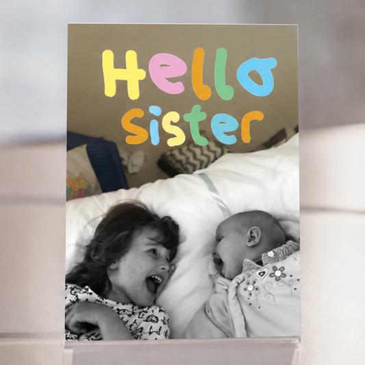 A portrait greeting card with the words 'Hello sister' in colorful playful lettering above a black-and-white photo of a young girl smiling at a baby.