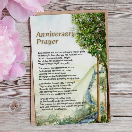 An 'Anniversary Prayer' card featuring a scenic forest background and gold lettering, displayed on a rustic wooden surface next to pink flowers.