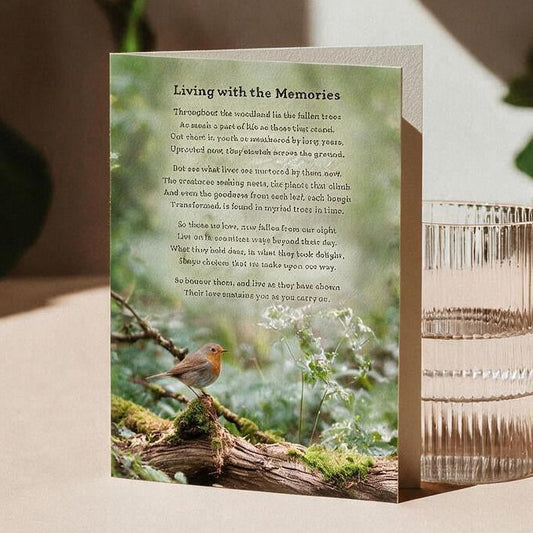 A portrait greeting card titled stands upright on a beige surface beside a ribbed clear glass tumbler. The front has a nature photograph of a small bird in a woodland with a poem printed below entitled "Living with the Memories".
