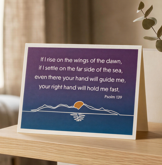 A Bible verse print card on a landscape card, placed on a light wood table in a softly lit room. The card has a purple-to-blue gradient background with white text and a simple mountain-and-sun illustration near the bottom.