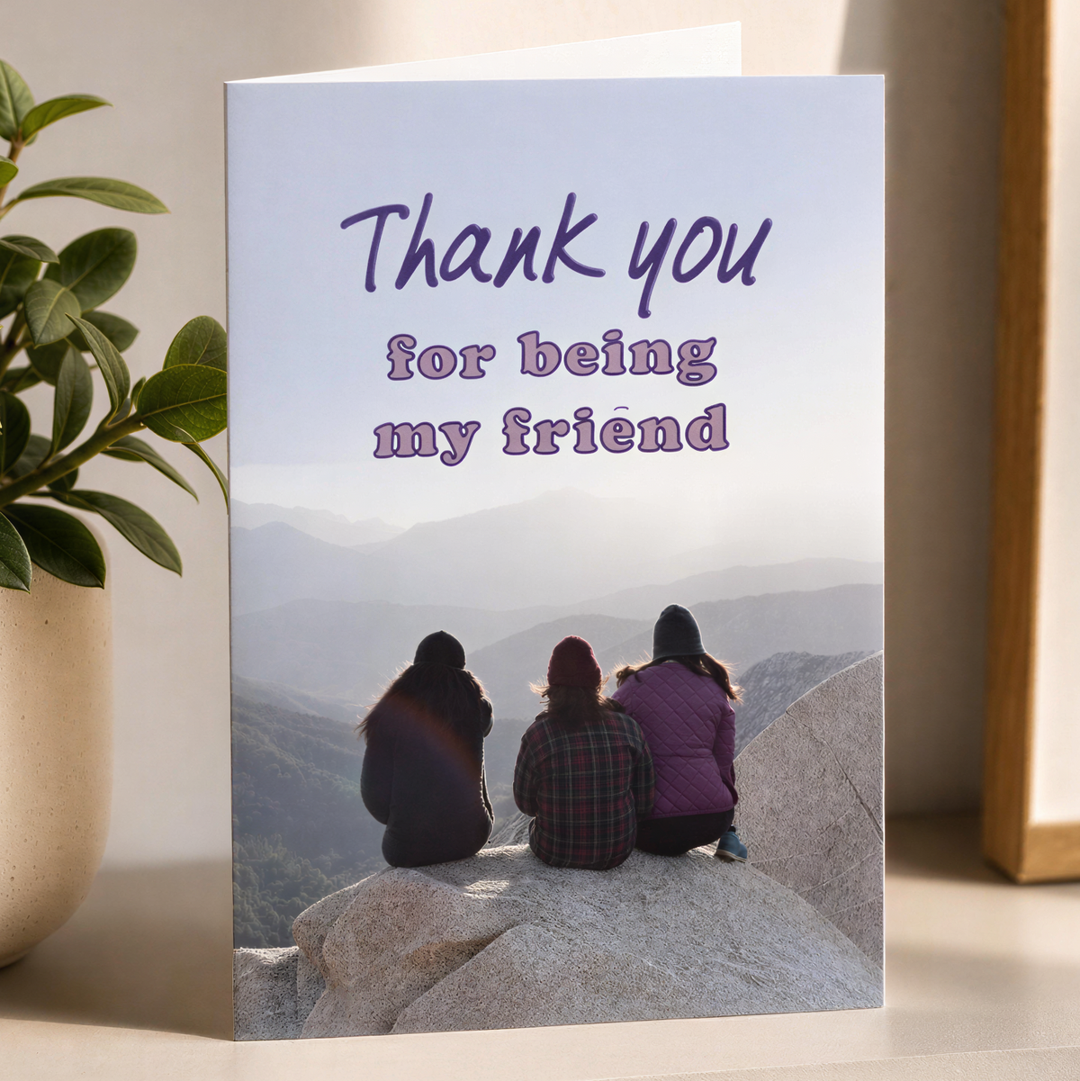 A portrait thank-you card standing on a white shelf, printed with the text 'Thank you for being my friend' above a photo of three people sitting on rocks overlooking a mountain landscape. A potted plant is on the left and part of a framed print is on the right, all against a warm beige wall.